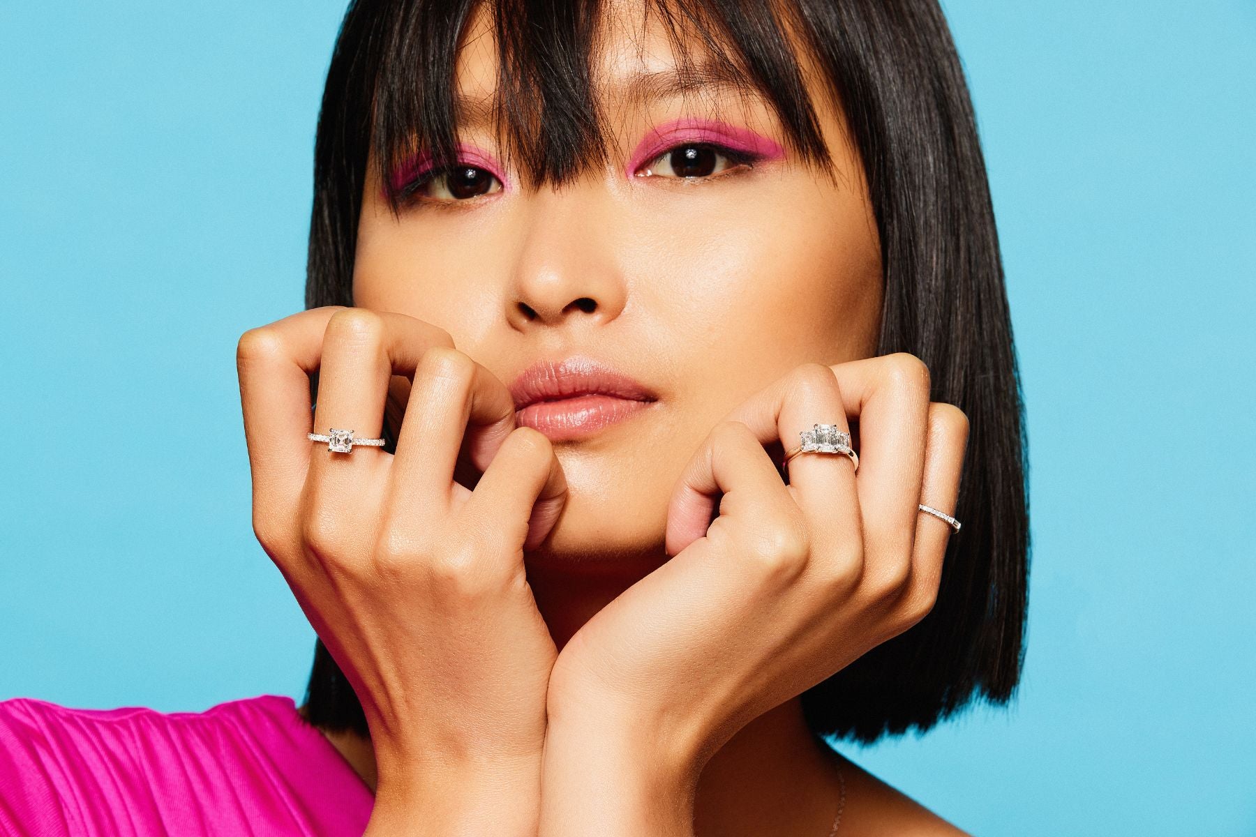 A person with straight black hair and bright magenta eye makeup poses against a blue background. They wear a pink top and display rings by placing their hands under their chin, highlighting the jewelry.