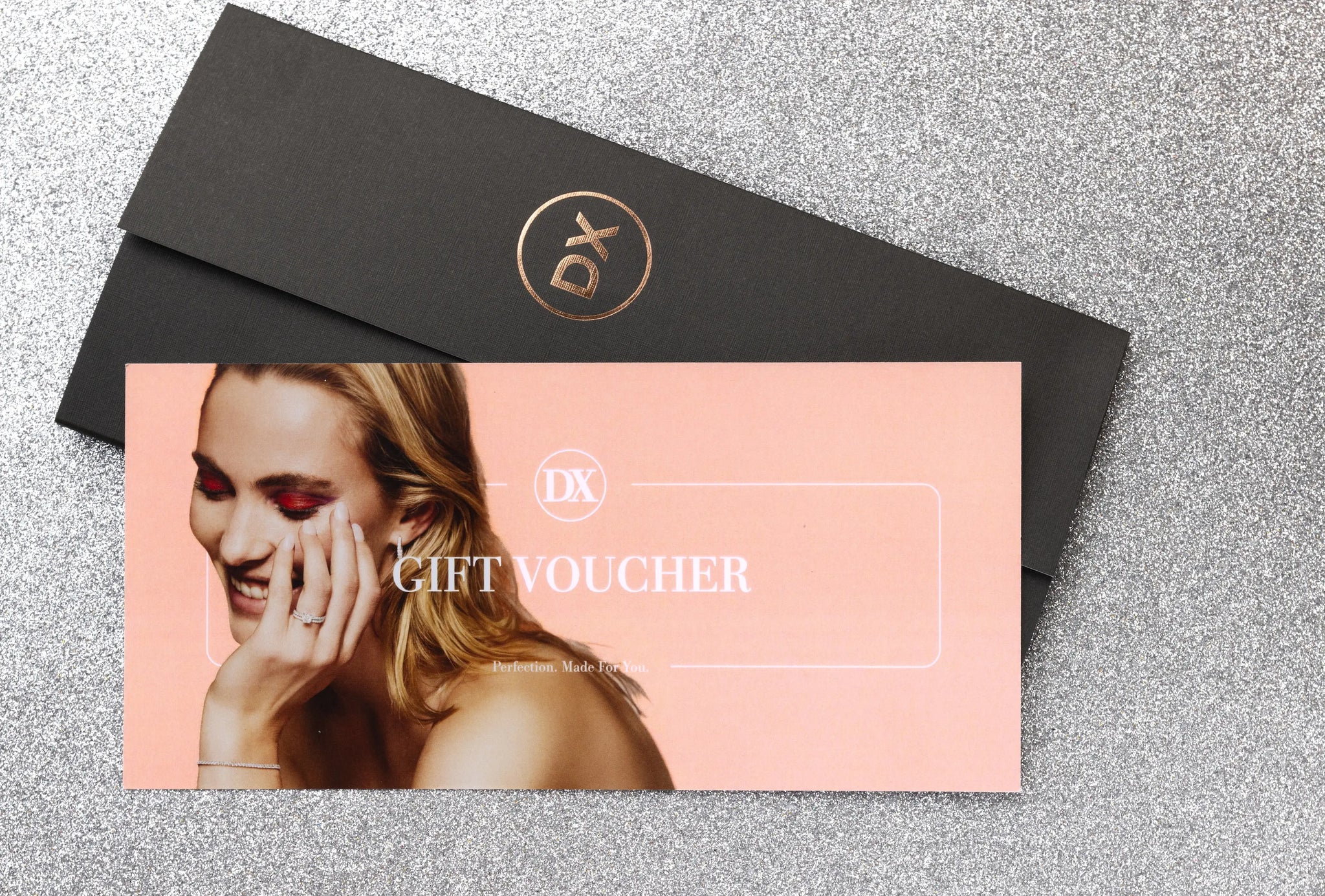 A DX Diamonds black envelope comes with a pink Gift Voucher, showcasing a joyful woman resting her hand on her face, hinting at the happiness brought by fine jewelry or an exquisite engagement ring.